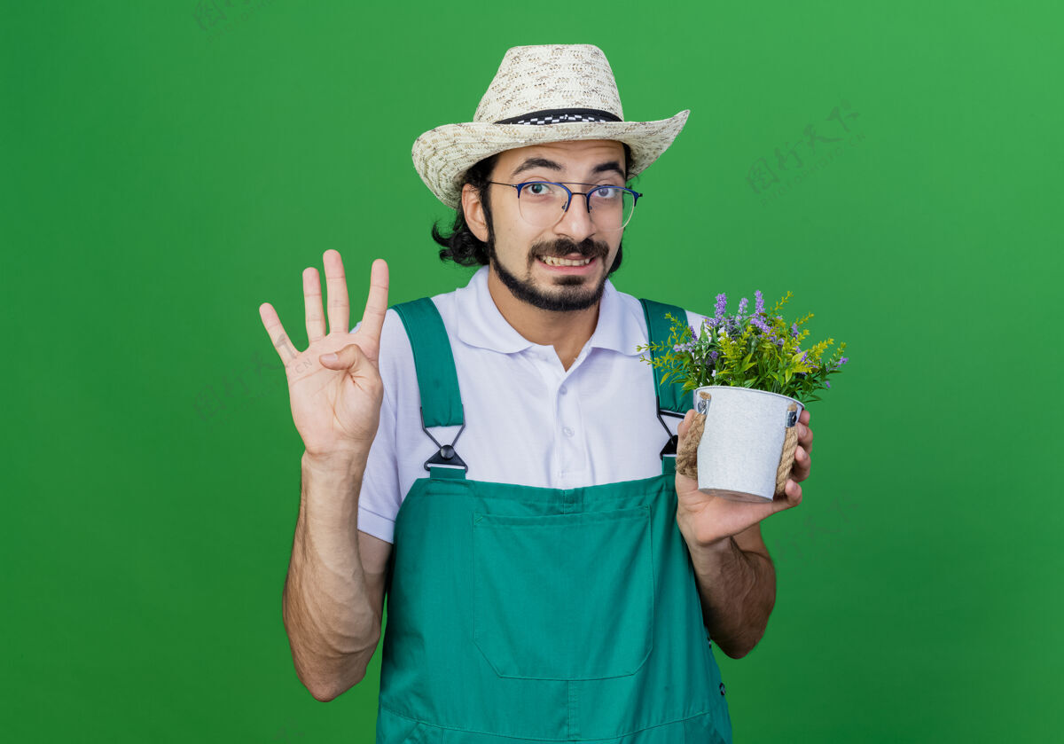 绿色年轻的留着胡须的园丁 穿着连体衣 戴着帽子 手里拿着盆栽植物 上面写着四号盆栽站着男人