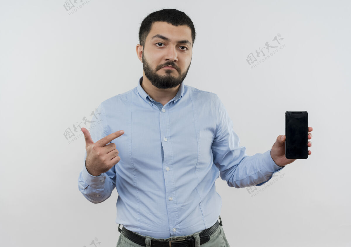 衬衫身穿蓝色衬衫 留着胡须的年轻人拿着智能手机 用食指指着它 感到困惑拿着男人留着胡子