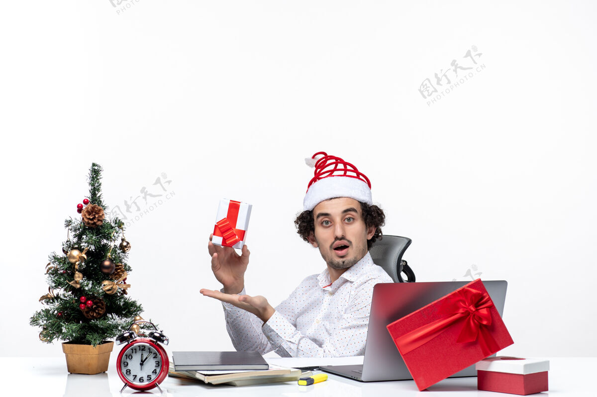 抱着带着圣诞老人帽子的年轻商人坐在办公室里 拿着礼物 在白色背景上摆姿势拍照 这让人感到很惊讶坐着帽子相机