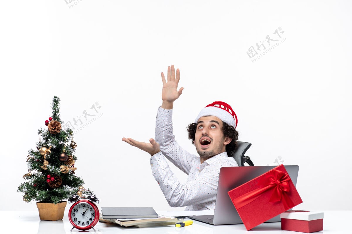 帽子带着微笑的新年心情积极的年轻商人戴着圣诞老人的帽子坐在办公室里 独自一人在白色背景下工作圣诞老人商人独自
