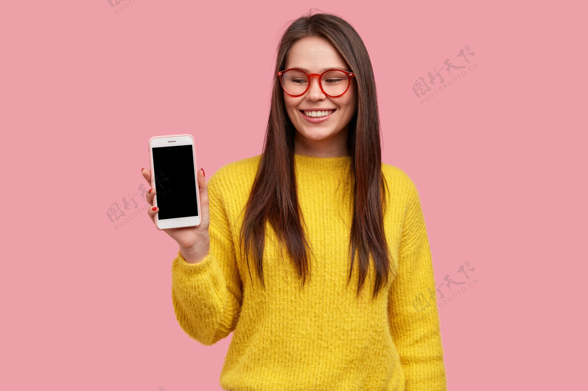 示范乐观的黑发女士展示智能手机屏幕 乐于购买新产品 戴眼镜和黄色毛衣电话展示保持