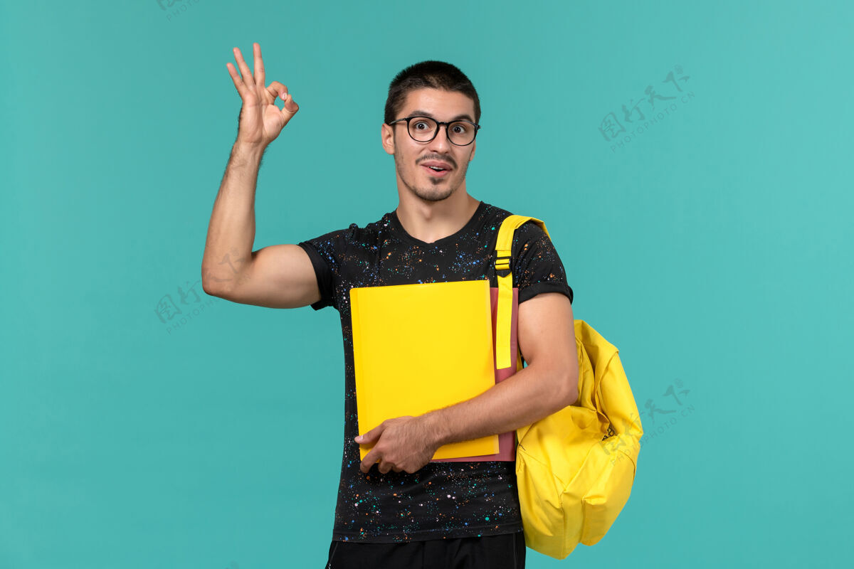 档案身穿深色t恤黄色背包的男学生正面图 浅蓝色墙上放着不同的文件人光大学