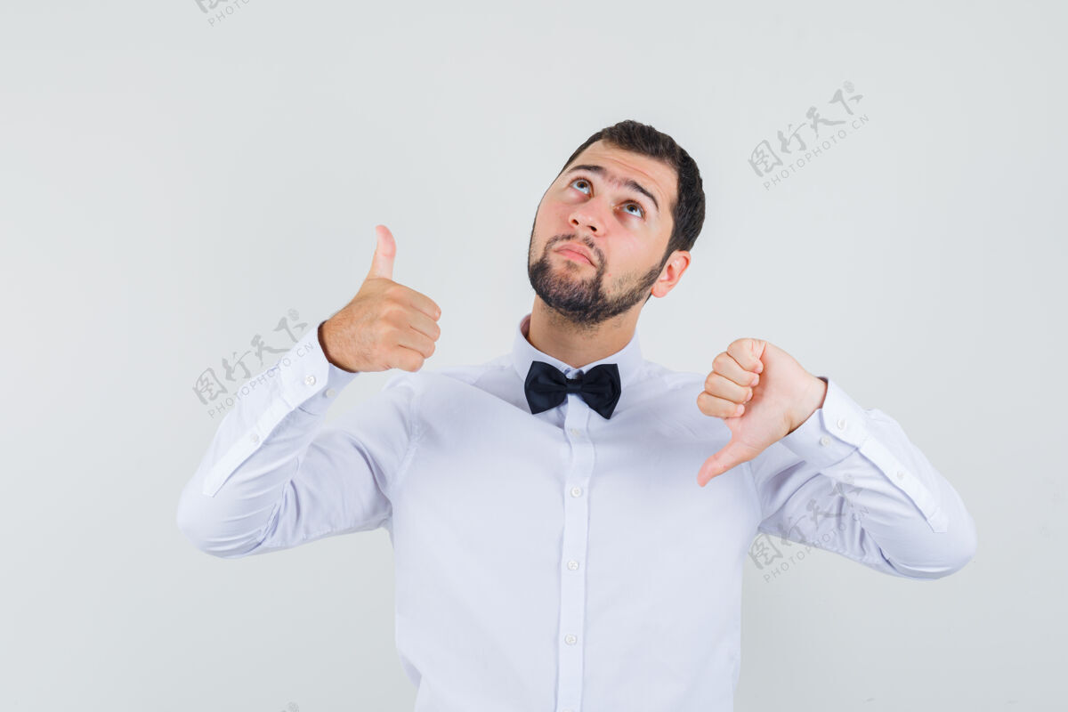 自信年轻的男性穿着白衬衫上下竖起大拇指 看起来很困惑 正面视图时尚站立成人