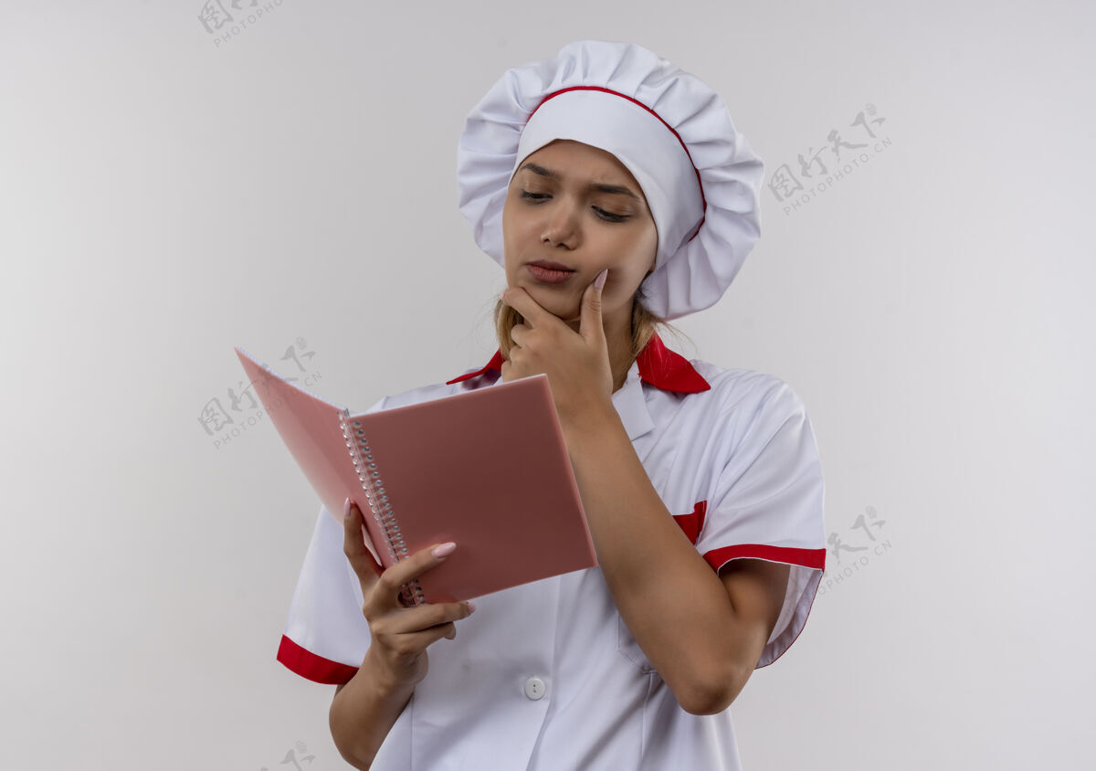 下巴困惑的年轻厨师穿着厨师制服的女厨师看着手里的笔记本 把手放在孤立的白墙上的下巴上放女穿