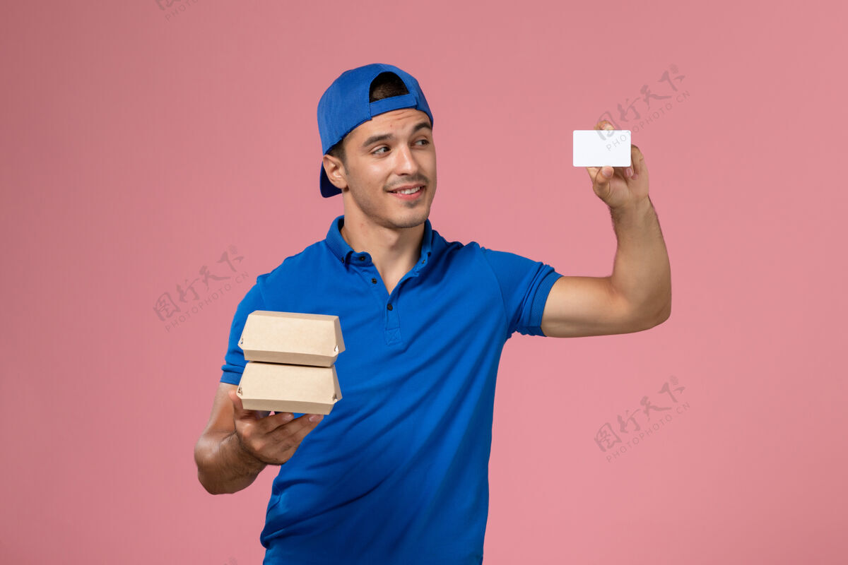食物正面图：身穿蓝色制服披肩的年轻男性快递员拿着小包裹 浅粉色墙上有卡片正面人持有