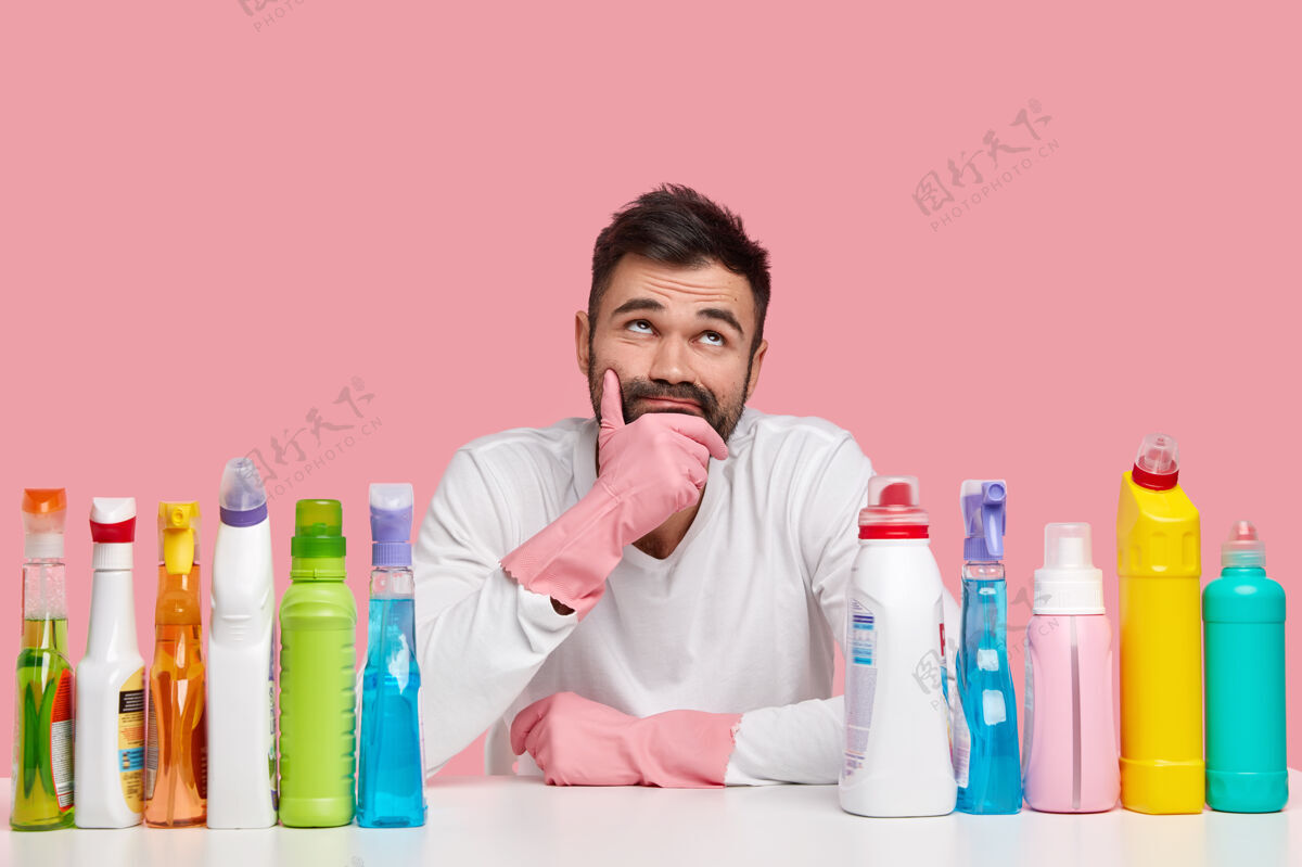 家务体贴男人的照片托着下巴 沉思地向上看 穿着白色的套头衫和手套 用洗涤液 清洁剂 隔离在粉红色的空间向上手套懒惰