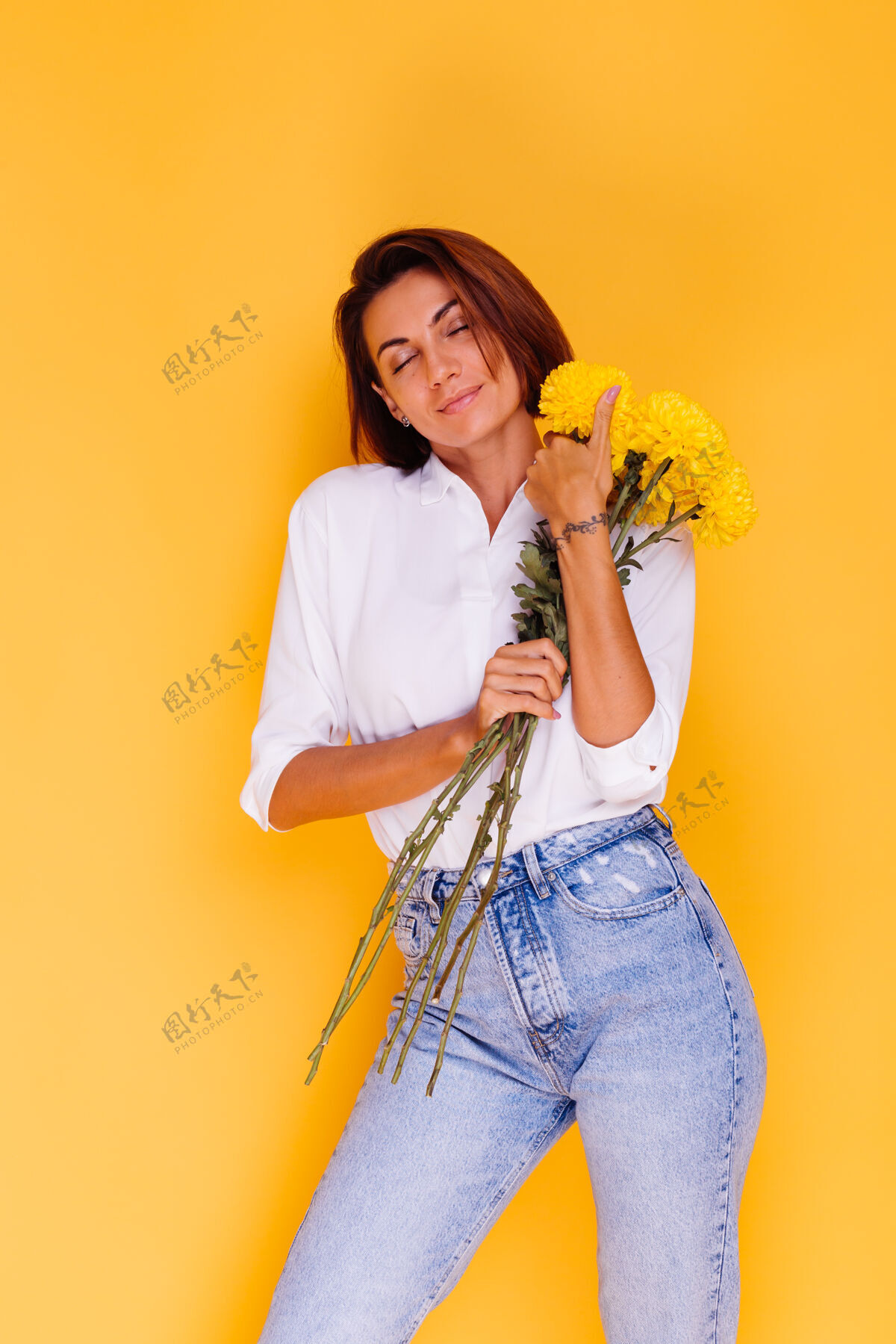 可爱摄影棚拍摄的黄色背景快乐的白人妇女短发穿着休闲服白衬衫和牛仔裤手持一束黄色紫苑微笑年轻惊喜