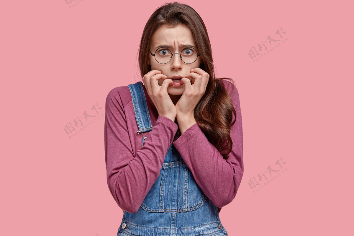 眼镜惊慌失措的关心的女人咬着手上的指甲 看起来紧张和害怕 戴着圆眼镜 牛仔工作服 隔离在粉红色的空间眼镜眼镜表情