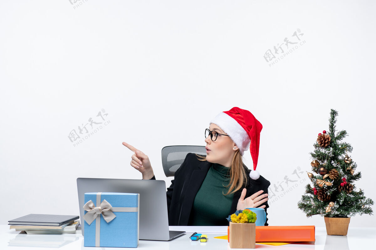 圣诞树一位商务女士惊讶地戴着圣诞老人帽坐在一张桌子旁 桌上放着圣诞树和礼物 她仔细地盯着白色背景上的东西圣诞老人圣诞节商务