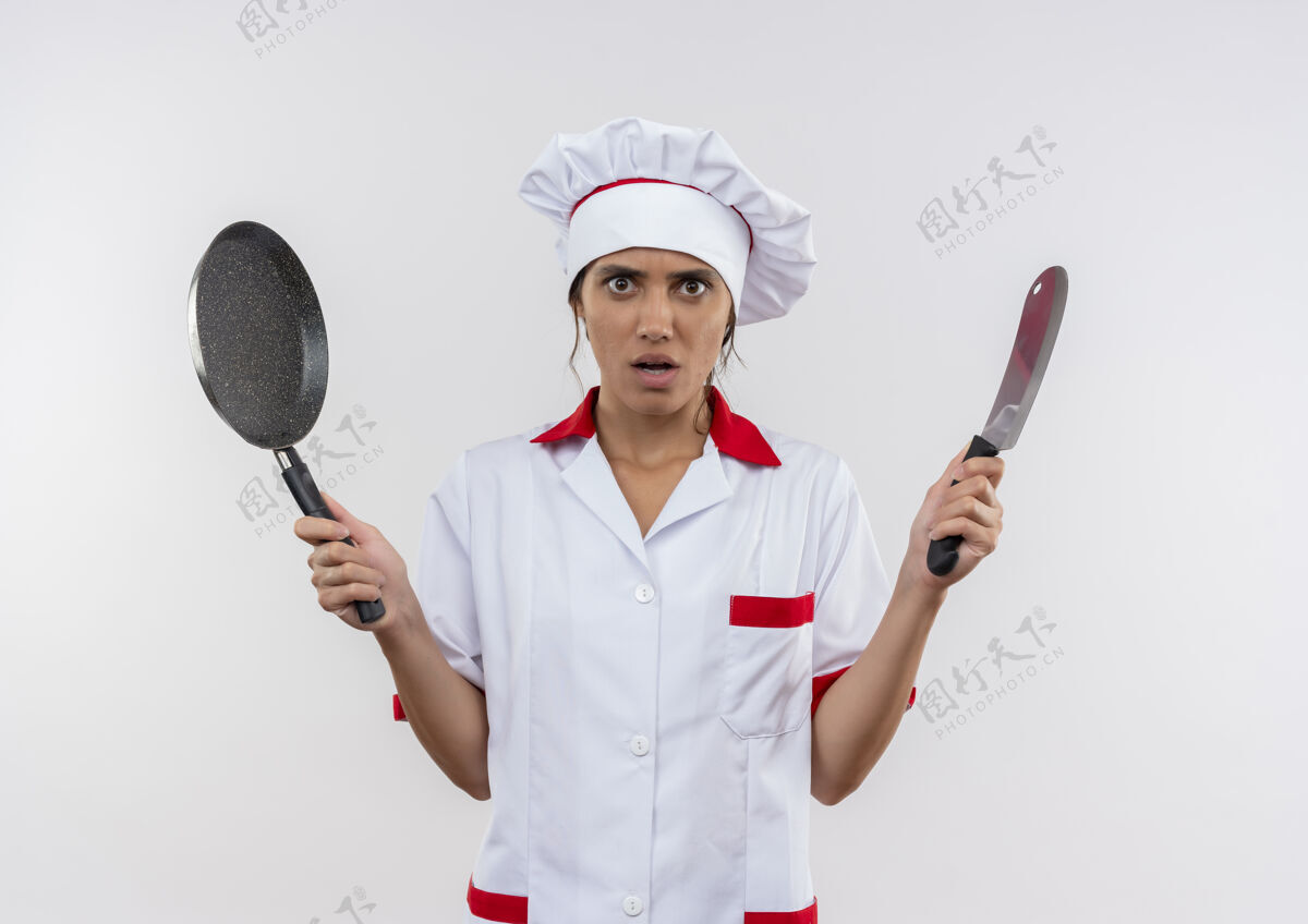 年轻年轻的女厨师穿着厨师制服 拿着菜刀和煎锅 站在隔离的白色墙壁上 留着复印空间平底锅拿着厨师