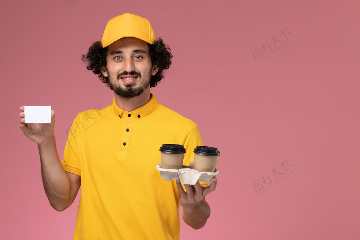 快递员正面图：身穿黄色制服和披风的男性信使手持棕色咖啡杯和粉色墙上的卡片棕色制服工人