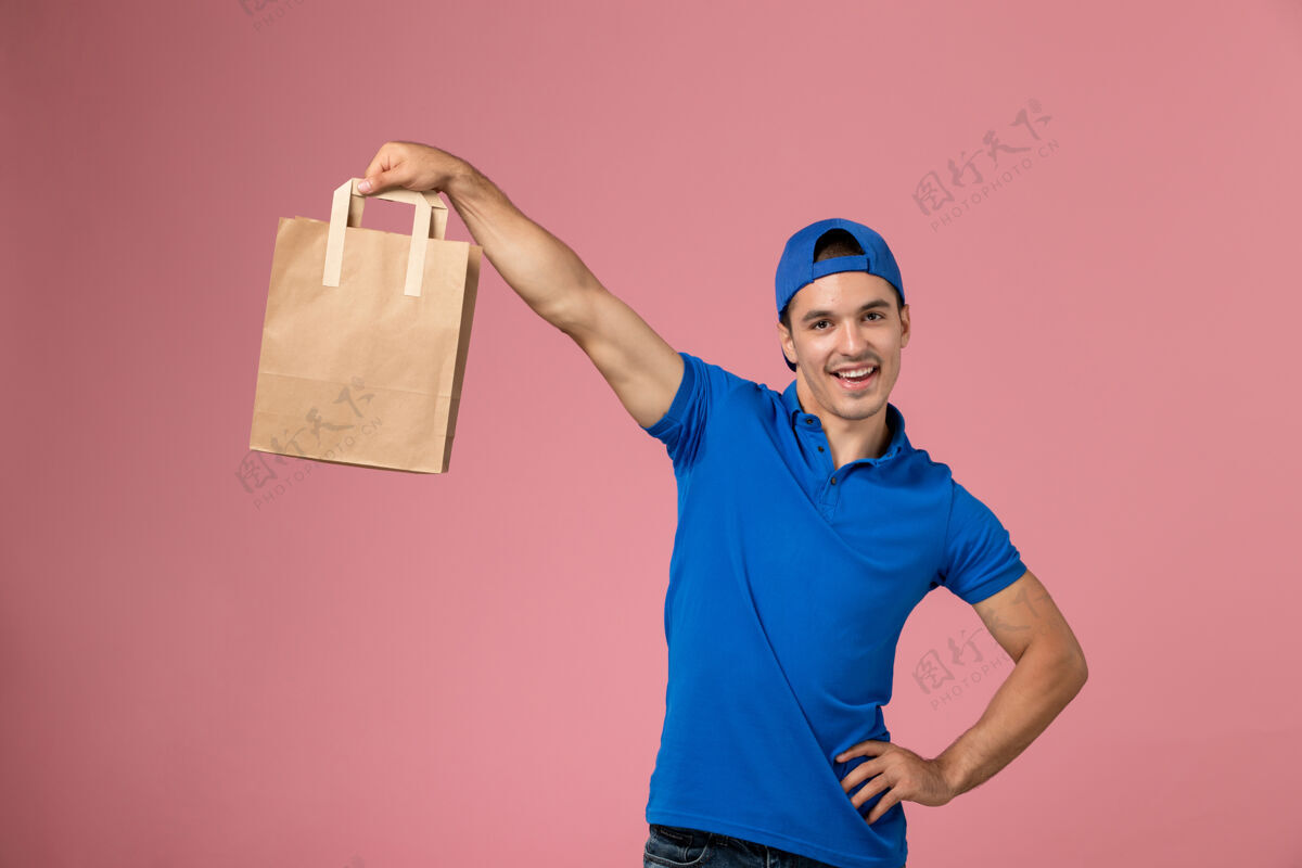 快乐正面图：身穿蓝色制服和斗篷的年轻男性快递员 手上拿着快递纸包裹 站在粉红色的墙上男人快递制服
